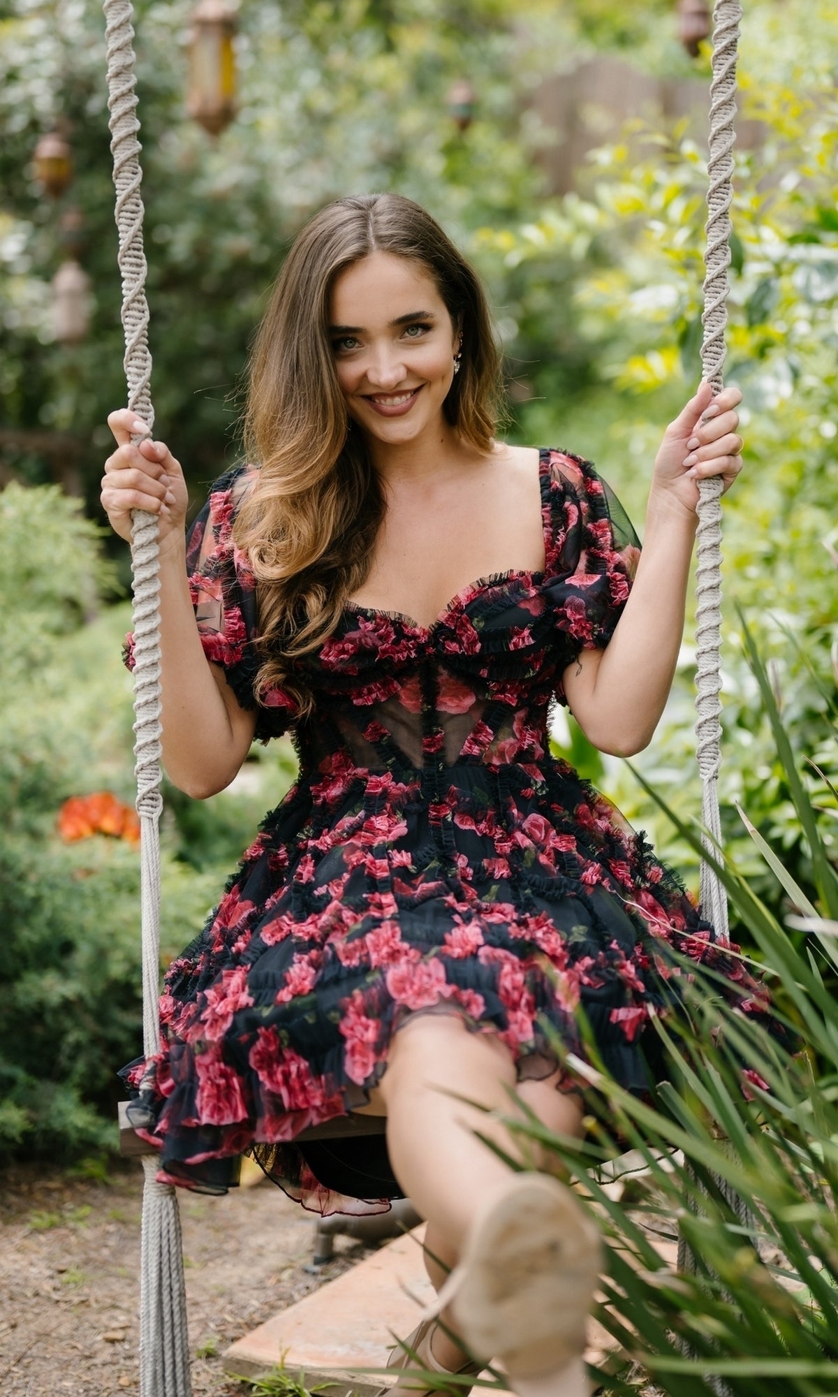 Wearing the Aleta 1304 Short Sleeve Short Print Prom Dress, a woman sits on an outdoor swing surrounded by lush green plants and flowers, smiling at the camera.
